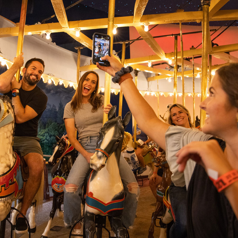 The grown-up on the right is turned towards two adults on the left. She is holding up her phone to take a picture of them riding horses on the Carousel.